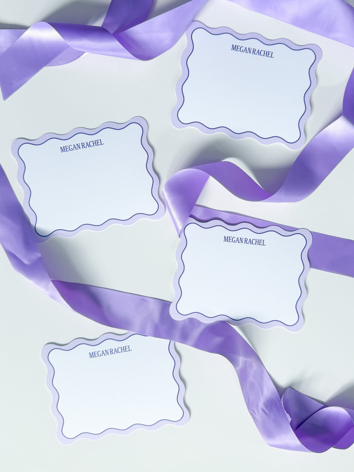 Five white paper notecards with lavender scalloped borders and the printed name 'MEGAN RACHEL' at the top, arranged on a white surface with flowing lavender ribbon.
